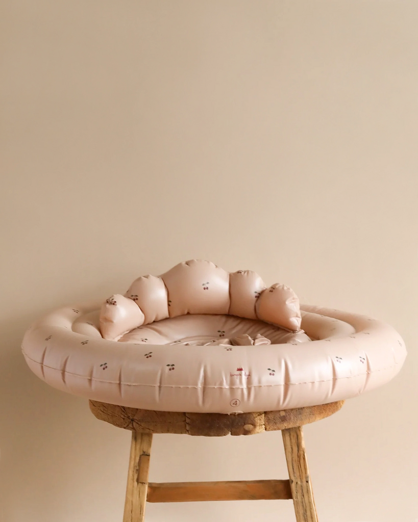 A soft, beige Inflatable Baby Swim Ring - Blush Cherry made from durable PVC features a cloud-like backrest and small printed patterns, ideal for ages 1-2 years. It rests on a wooden chair against a plain beige background, resembling the comfort of an inflatable swim ring.