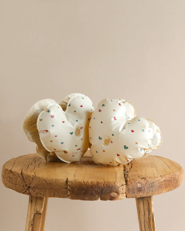 Two cream-colored, heart-shaped cushions with scattered small, multi-colored hearts lie on a rustic wooden stool. The cushions are slightly shiny and have a soft, puffy appearance, creating a cozy and cute aesthetic. They almost look like adorable Inflatable Water Wings - Multi Hearts amidst the neutral beige background.