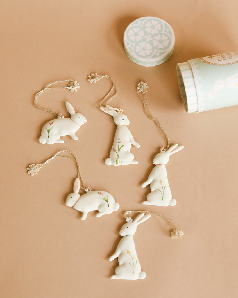 Five sweet Maileg Easter Bunny Ornaments, 5 pcs. with floral details are spread out on a beige surface. Each is attached to a string with a flower-shaped bead. A cylindrical container decorated with similar rabbit designs is partially opened, lying to the right of these Easter decorations.
