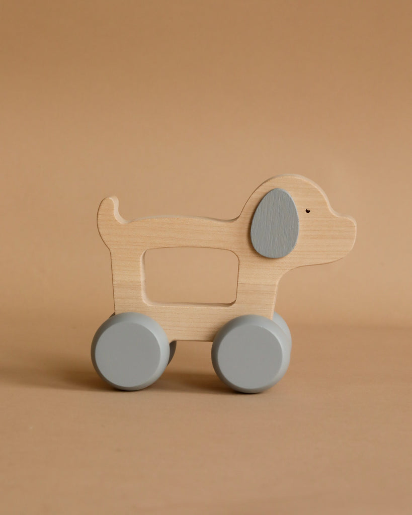 A Wooden Puppy Push Toy is set against a beige background. Made in Poland, the toy features light-colored wood with gray accents on the ear and wheels. The simple and minimalist design aids in developing fine motor skills, making it ideal for young children.