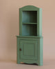 The Maileg Large Corner Cabinet - Dark Green is a petite wooden piece featuring a closed door at the bottom and an open shelving unit above it. The shelves are empty, and the cabinet is set against a plain beige background. Its curved top, vintage finish, and simple door knob bring stylish home decoration charm to any room.