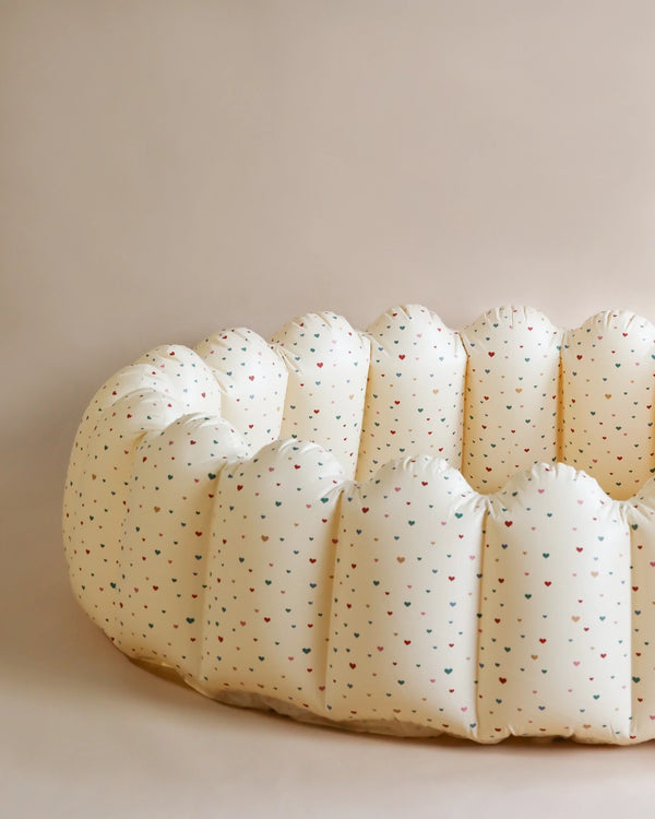 A close-up of a soft, round, beige cushion with small, colorful heart-shaped patterns. The Inflatable Large Shell Pool - Multi Hearts, resembling an inflatable pool float, has a tufted appearance and is set against a plain, light beige background.