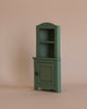 The Maileg Corner Cabinet, Mouse in Dark Green features a vintage finish, with a single closed door and two open shelves above it. Its simple, miniature wooden design fits perfectly against the plain beige background, making it an ideal piece for home decoration.