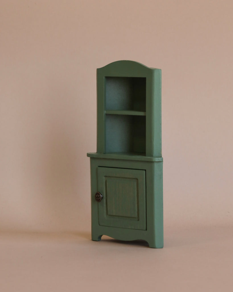 The Maileg Corner Cabinet, Mouse in Dark Green features a vintage finish, with a single closed door and two open shelves above it. Its simple, miniature wooden design fits perfectly against the plain beige background, making it an ideal piece for home decoration.