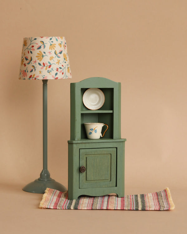 The Maileg Corner Cabinet, Mouse - Dark Green, with a vintage finish, features a small cabinet door below and two shelves above holding a floral teacup and a white plate. It stands on a colorful striped rug beside a tall, thin lamp with a floral-patterned lampshade, enhancing the home décor against the plain beige wall.
