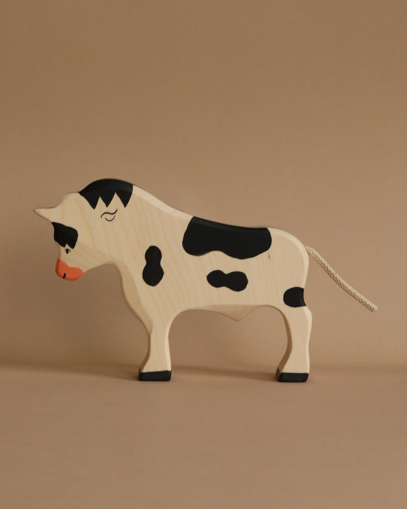 A wooden Holztiger Bull with black and white spots stands against a neutral brown background. Handcrafted wood brings out its simple, stylized design with a short tail and slightly lowered head. This charming figure is part of the HOLZTIGER collection, proudly Made in Europe.