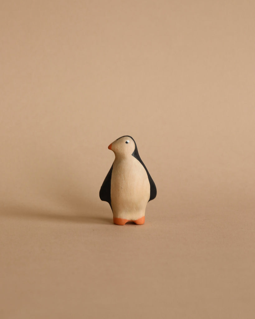 A Handmade Wooden Penguin stands against a warm beige background. Handcrafted from linden wood and made in Serbia, the figurine is stylized with a simple black and white body and orange feet, all painted with non-toxic child-safe paint.