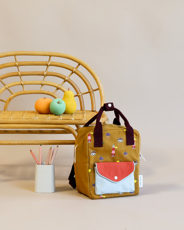 A Sticky Lemon Backpack Small Special Edition | Adventure Collection | Khaki Green made of waterproof nylon with a red flap pocket, adorned with playful patterns, sits on the floor next to a bamboo chair. On the chair are three colorful fruit-shaped objects, and a white container on the floor holds several colored pencils.