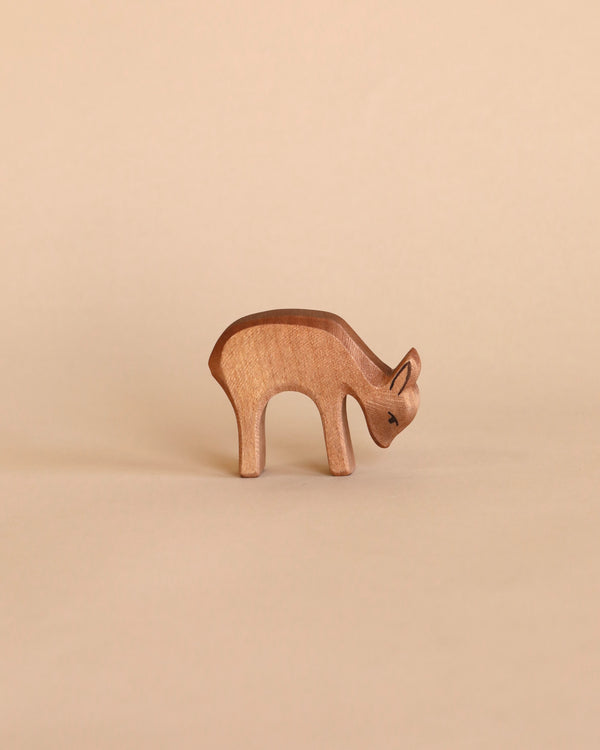 A handcrafted wooden figure of an Ostheimer Deer - Eating, carved with simplistic details and smooth edges, stands against a plain, light beige background.