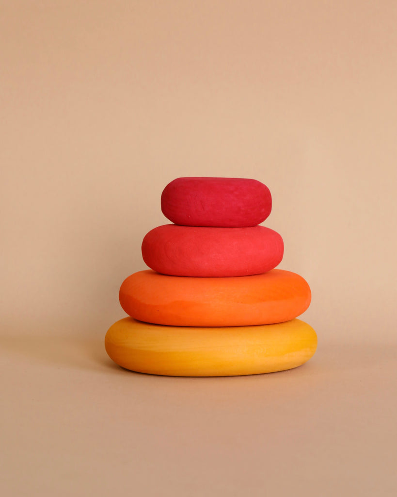 A stack of four smooth, flat stones is arranged in decreasing size from bottom to top. The stones, shaped organically and vibrantly colored in yellow, orange, light red, and dark red using non-toxic color stains, are set against a neutral beige background. This beautiful arrangement is known as the Grimm's Fire Pebbles Stacker.