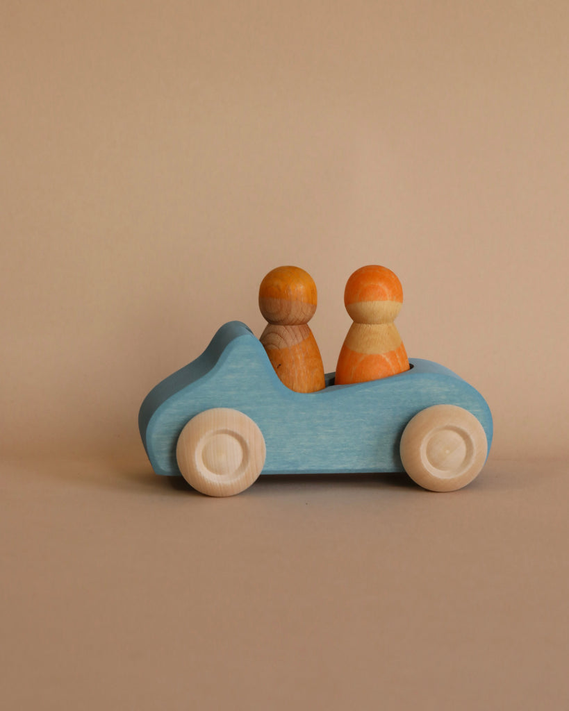 A Grimm's Wooden Blue Convertible Car painted light blue with rounded wheels and two simplistic wooden figurines as passengers, one colored orange and the other natural wood. The toy features a non-toxic water-based stain, set against a beige background.