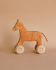 A Grimm's Wooden Horse Willy Push Toy with simple features and four white wheels stands against a neutral beige background. Made in Germany, the push toy has a smooth finish, a small visible eye, and a brown tail made of a thin strip of non-toxic material.