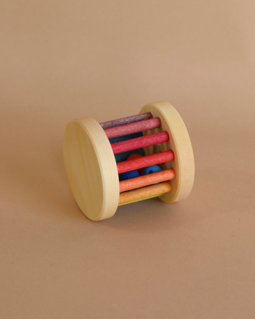 A Mini Rolling Wheel with a cylindrical shape, featuring two flat circular sides and colorful rainbow bars connecting them. Inside the rattle, a small blue ball is visible between the bars, resembling a mini rolling wheel. The background is a plain, light beige surface.