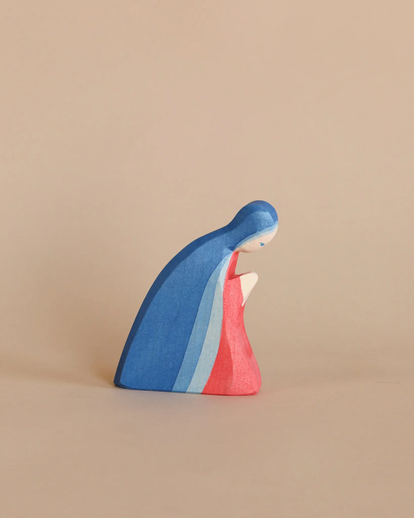 The Ostheimer Nativity Scene features a handcrafted wooden figure of a person in profile, draped in a cape of blue, light blue, and pink. The figure is depicted seated with their head slightly bowed on a beige background and is crafted from sustainably sourced materials.