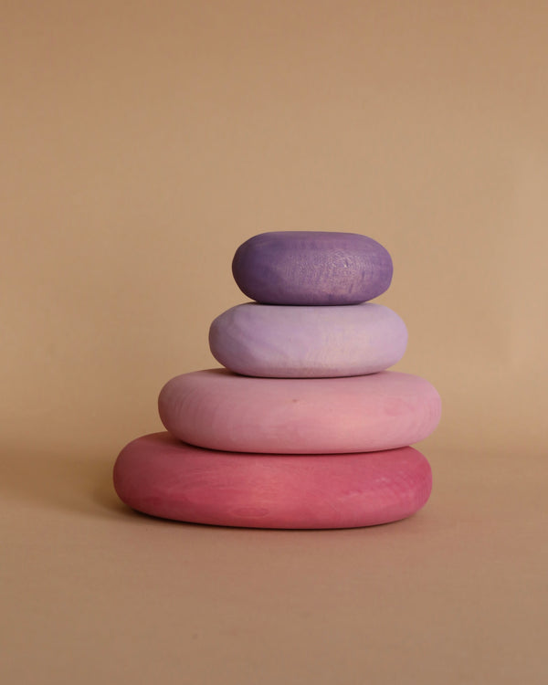 A stack of four smooth, rounded stone-like objects in varying shades of purple and pink is seen against a beige background. The non-toxic objects are arranged from largest at the base to smallest at the top, building a visually pleasing gradient. These are the Grimm's Flamingo Pebbles Stacker.