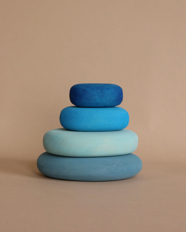 Four smooth, flat, organic stone shapes are stacked on top of each other against a beige background. The stones are in shades of blue, with the largest stone at the base in light blue and the smallest stone at the top in a darker blue. This is the River Pebbles Stacker.