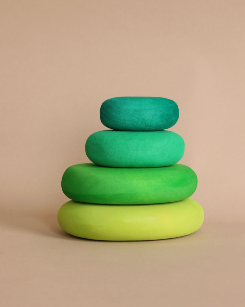 Four stacked, smooth, pastel-colored stones in varying shades of green sit on a light beige background. Resembling organic stone shapes, the Moss Pebbles Stacker decreases in size from bottom to top, creating a balanced, gradient effect that's reminiscent of building and stacking toys.