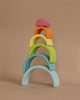 A stack of Grimm's Small Wooden Rainbow, Pastel arches in various colors with the smallest, pink arch on top, followed by orange, yellow, green, and blue arches—all crafted from non-toxic wood—each increasing in size from top to bottom. This versatile toy sits against a neutral beige background.