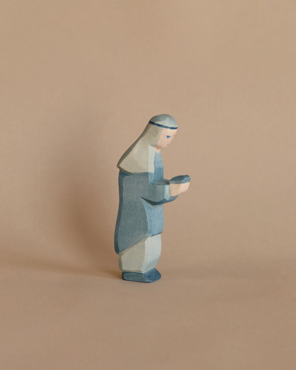 A small, handcrafted wooden figurine, known as the Ostheimer Blue King, features a character adorned in a blue robe with a head covering, holding a small object in their hands. The background is a plain, light brown surface, making it perfect for imaginative play with other Ostheimer toys.