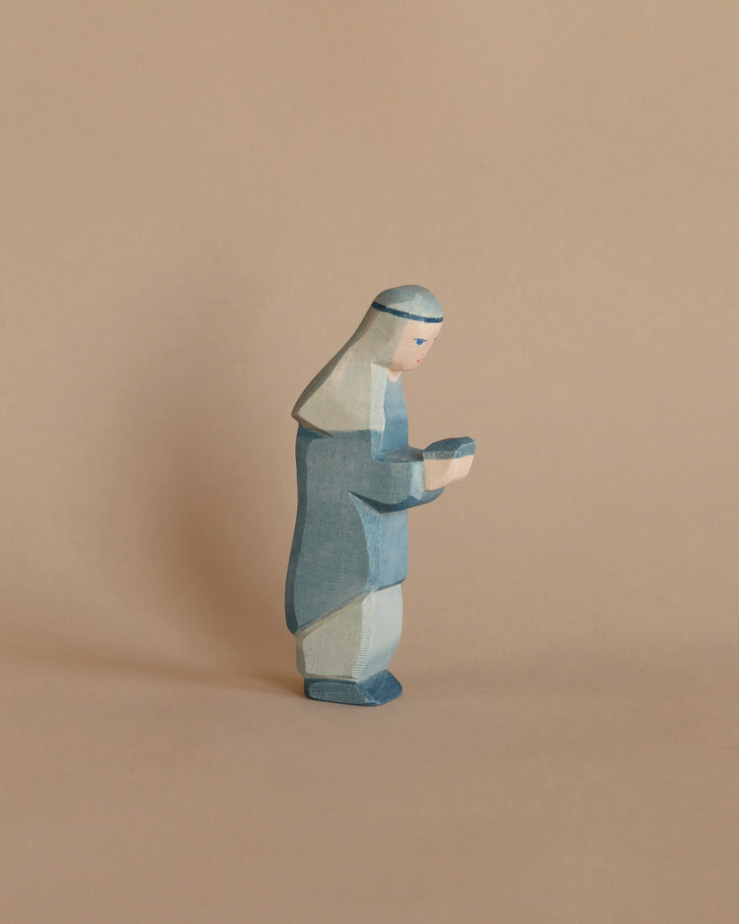 A small, handcrafted wooden figurine, known as the Ostheimer Blue King, features a character adorned in a blue robe with a head covering, holding a small object in their hands. The background is a plain, light brown surface, making it perfect for imaginative play with other Ostheimer toys.