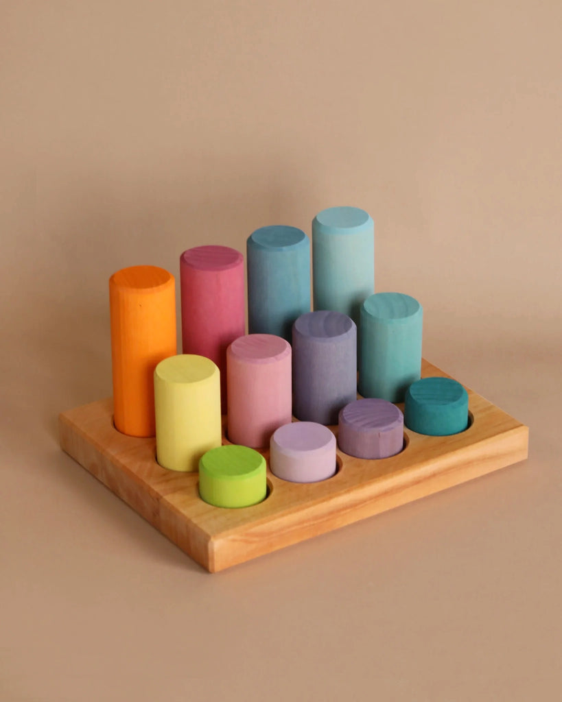 A wooden board holds 12 cylindrical blocks painted in various pastel colors. The cylinders, resembling construction pieces, are arranged in rows with the tallest at the back and the shortest at the front, creating a stepped effect. Ideal for enhancing motor skills, this Grimm's Stacking Game Small Pastel Rollers sits on a plain beige surface.