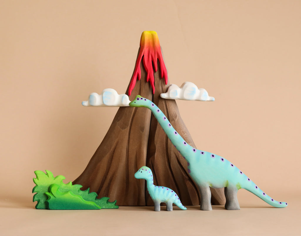 A Handmade Wooden Volcano & Dinosaurs Set featuring a volcano with red and yellow lava, two clouds, a large green dinosaur, a small green dinosaur, and a leafy bush. The background is beige, enhancing the wooden textures and colorful accents of the non-toxic paint used on these charming toys from Romania.