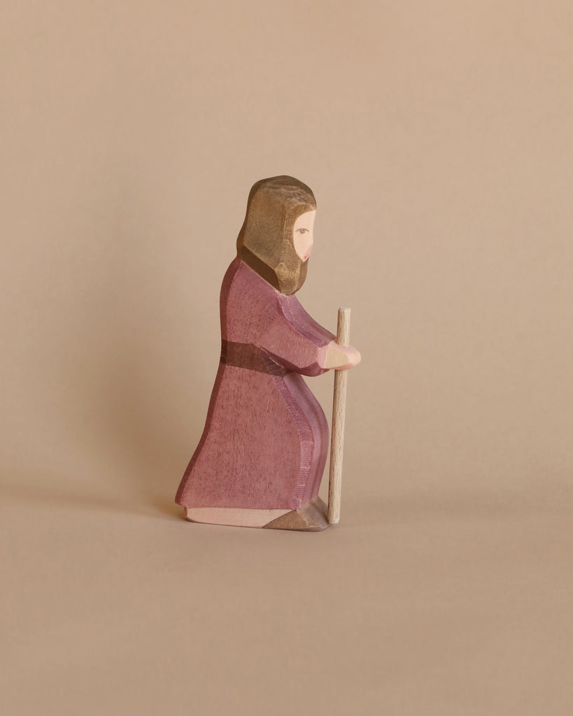 A handcrafted Ostheimer Joseph - Standing figurine of a bearded man in a purple robe and hood, kneeling and holding a staff, set against a plain beige background.