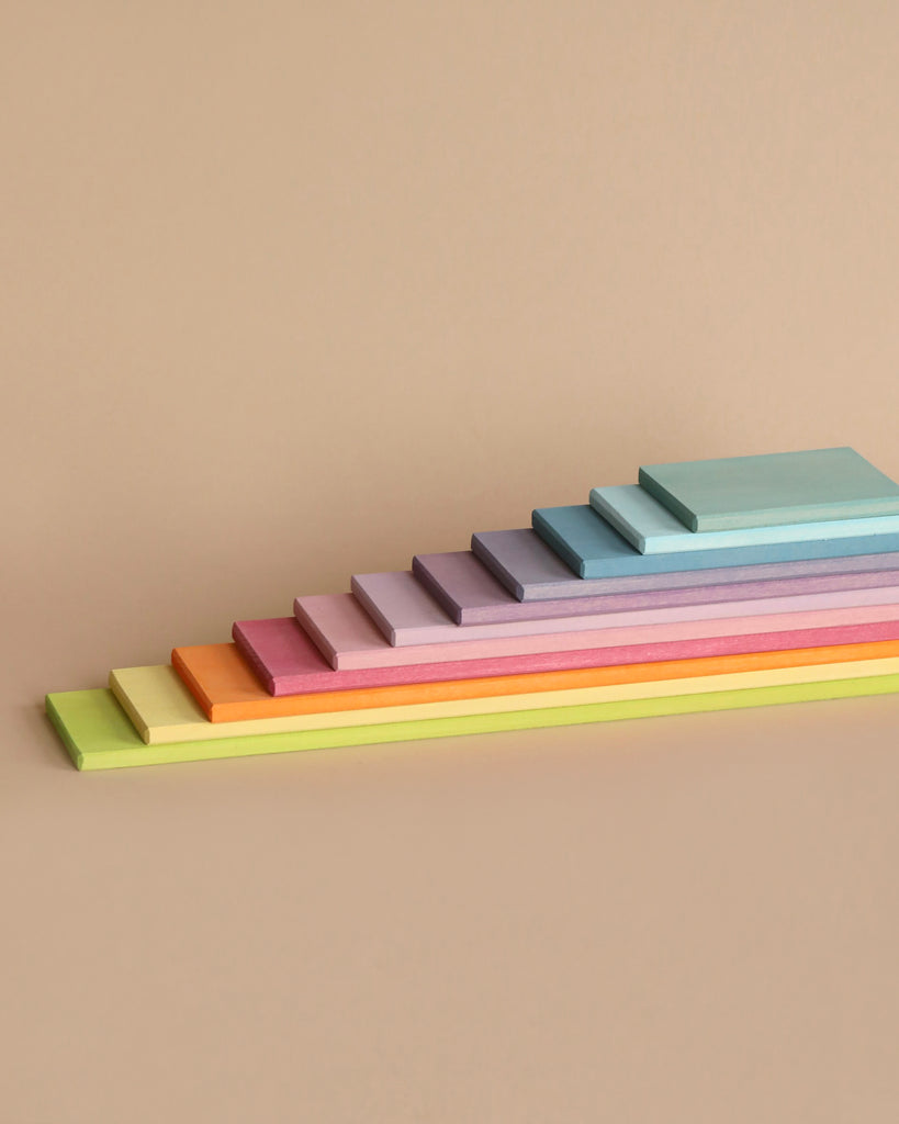 A neat arrangement of Grimm's Pastel Building Boards Set in ten different colors, each one slightly larger than the one before it, creating a stair-step effect. Colors include green, yellow, orange, pink, purple, blue, and others—perfect for open ended play—displayed against a beige background.