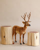 A Large Deer Stuffed Animal - Male (final sale) stands between two wooden stump stools. The background is a plain, light beige color, adding to the natural, rustic appearance of this plush animal setting.