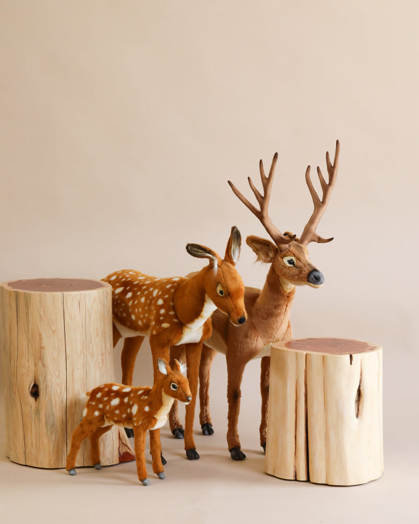 A group of three Large Deer Stuffed Animal - Female toys are arranged among light-colored wooden logs. The plush animals include an adult male with antlers, an adult female, and a fawn, all with realistic features. The background is a simple, neutral beige color.