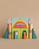 A Grimm's Building World Cloud Play Block Set made of solid wood blocks, including arches, cylindrical columns, and step-like pieces. Two wooden peg dolls are placed at the entrance—one in orange and another in white. The scene invites imaginative free building against a warm beige background.