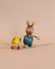 A cherished Collectible Dregeno Easter Figure - Rabbit With Trolley depicting a bunny with a beige and blue body pushing a small yellow toy chick on a red-wheeled cart, set against a plain light beige background.