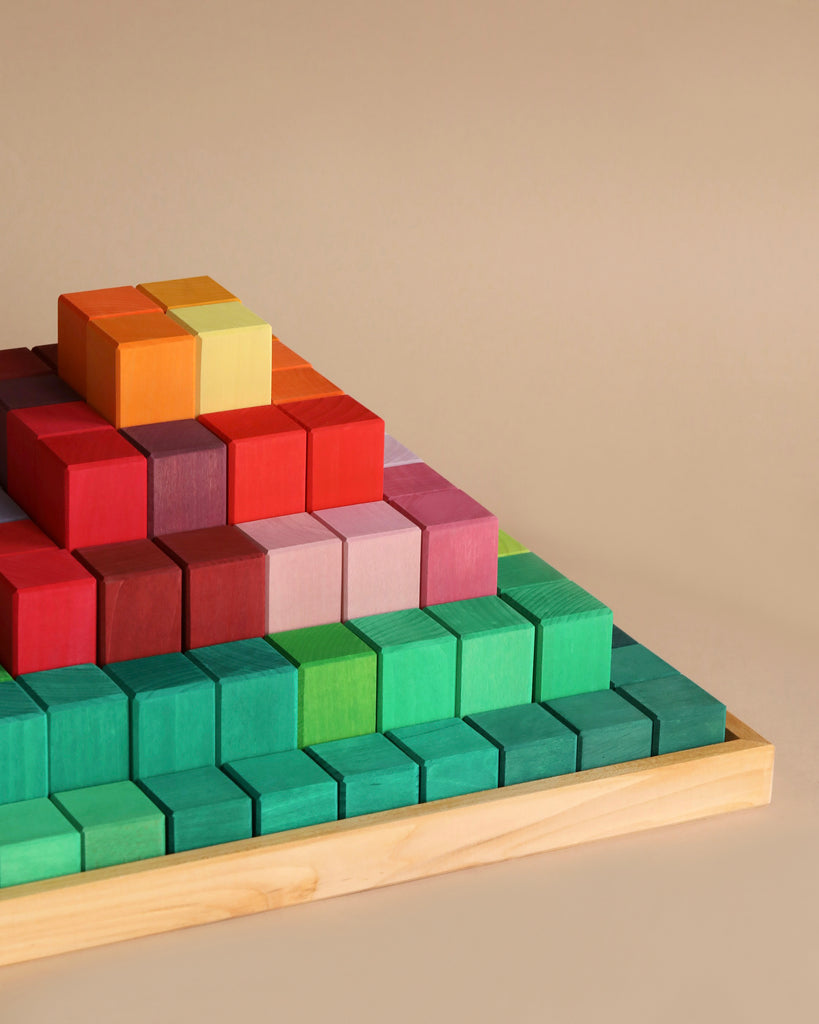 A Large Stepped Pyramid Block Set made of non-toxic wood blocks in various shades of green, teal, red, orange, yellow, and pink is arranged on a wooden tray. The 100 building blocks are stacked in layers, creating a tiered structure. The background is a neutral beige.