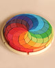 A Grimm's Wooden Color Spiral Block Set composed of interlocking, crescent-shaped pieces arranged in a rainbow gradient. The colors transition smoothly from red and orange to yellow, green, blue, and purple, creating a visually striking multicolored spiral pattern that enhances fine motor skills for young builders.