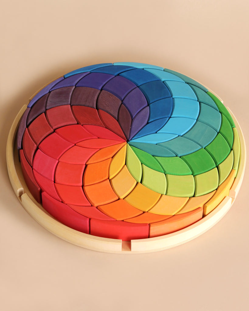 A Grimm's Wooden Color Spiral Block Set composed of interlocking, crescent-shaped pieces arranged in a rainbow gradient. The colors transition smoothly from red and orange to yellow, green, blue, and purple, creating a visually striking multicolored spiral pattern that enhances fine motor skills for young builders.