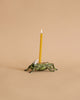 A whimsical candle holder shaped like a green frog, with a lit yellow birthday candle positioned on its back, set against a plain beige background.