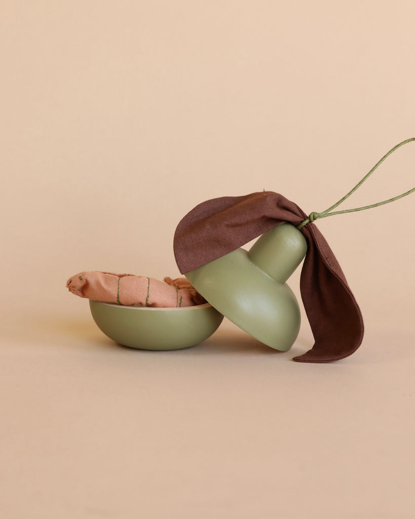 Product Description: The Wooden Pear & Little Friend set includes a minimalist green bowl adorned with a matching lid-like structure, topped elegantly with a dark brown fabric bow, giving it the appearance of a mushroom. Inside the bowl, you'll find an eco-friendly toy designed as a small piece of wooden fruit alongside the lid. The backdrop is a simple, light beige.