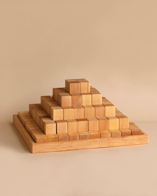 A large stepped pyramid-shaped structure made from 100 Grimm's Large Natural Stepped Pyramids. The blocks are arranged neatly in a square base, gradually decreasing in size as they stack upwards, forming a smooth, stepped pyramid. The background is a plain, light beige color.