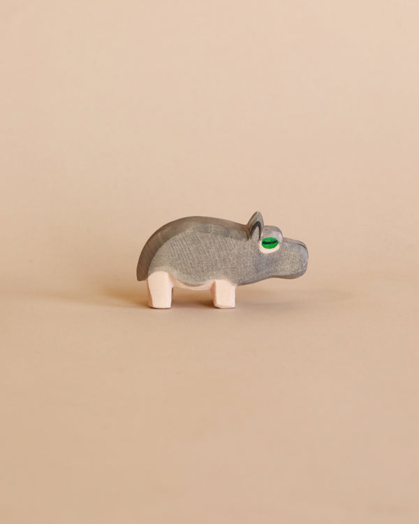 A small Ostheimer Hippopotamus figurine with visible wood grain, green eyes, and pale pink legs stands against a plain beige background.