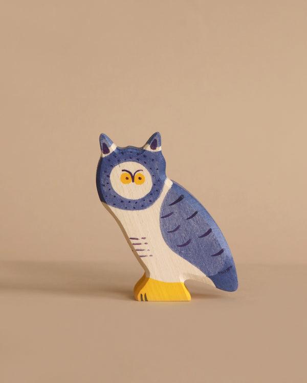 A small, wooden owl figurine is painted using watercolors in a mix of blue, white, and yellow tones. The owl features big, round yellow eyes, blue wings with darker blue markings, and yellow feet. The backdrop is a plain beige surface. This piece fits seamlessly among handcrafted wood toys like the Holztiger Owl.