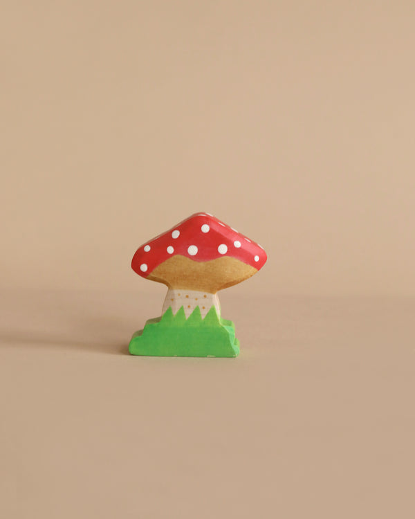 A small, handcrafted wooden mushroom figurine is centered against a beige background. The mushroom has a red cap with white spots and a light brown stem. It stands atop a green base resembling grass, reflecting the quality of Holztiger Toadstool figures crafted from maple and beech wood.