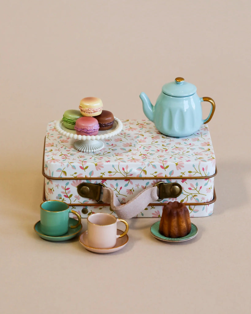 A delightful display featuring the Maileg Royal Tea Party includes a floral-patterned suitcase reminiscent of a Maileg Castle, acting as a table to support a blue teapot and vibrant macarons. Beneath it rest two cups with saucers and a small cake, portraying an enchanting, vintage tea party ambiance.