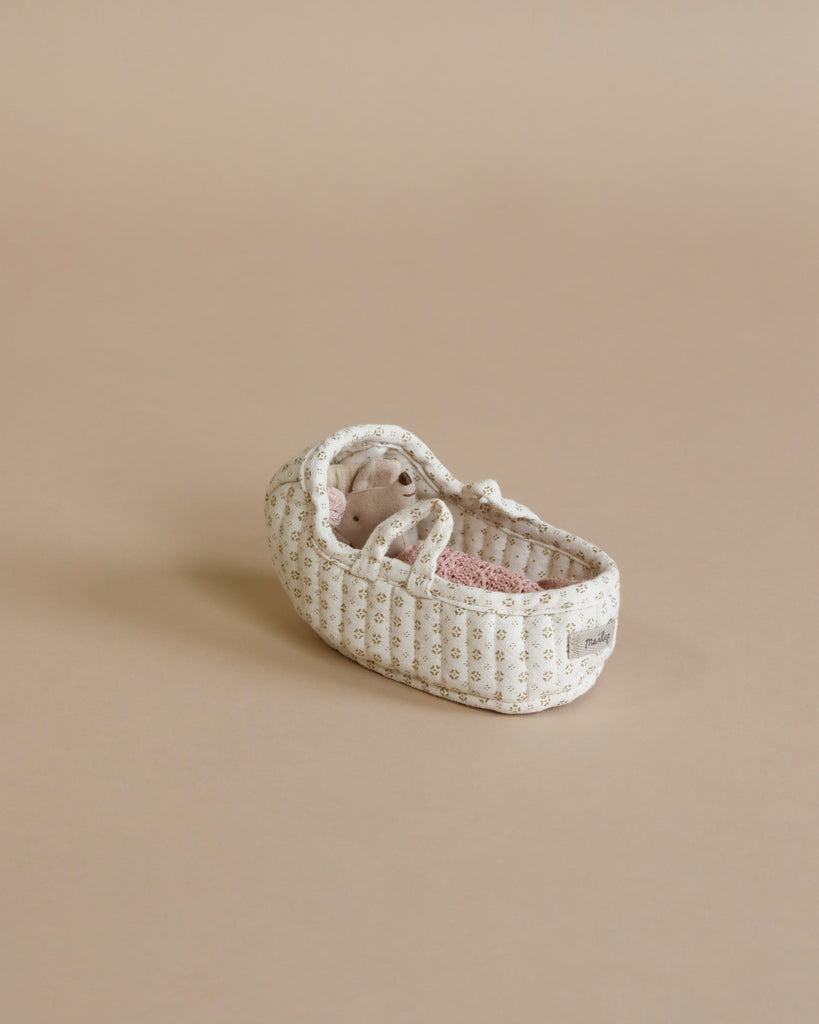 A small baby doll in a light beige patterned basket cradle with a pink mattress, laying on a neutral beige surface. The Maileg Carry Cot, Micro, designed like a micro sized bunny bed, has braided handles and the doll is nestled inside, appearing cozy and comfortable.