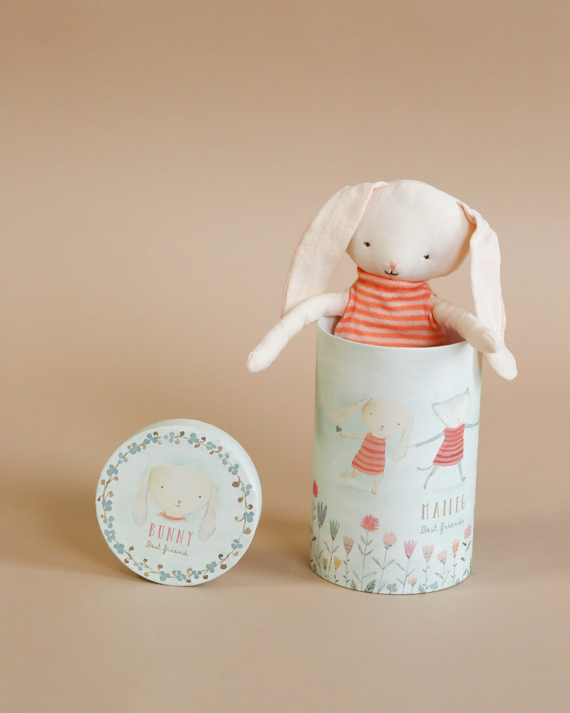 A Maileg Best Friend - Bunny wearing an orange striped shirt is peeking out of a gift tube adorned with illustrations of bunnies and flowers. The words "MALLIE" and "Best Friend Bunny" are printed on the container. A matching lid with the word "BUNNY" lies beside it, making it perfect for all ages.