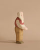 An Ostheimer Grandfather, a wooden figurine of a standing person with minimalistic features, handcrafted in the renowned Ostheimer tradition. The figure has a white head, a red shirt with a light pink arm, beige pants, and dark brown shoes against a plain beige background. The style is simplistic and blocky.