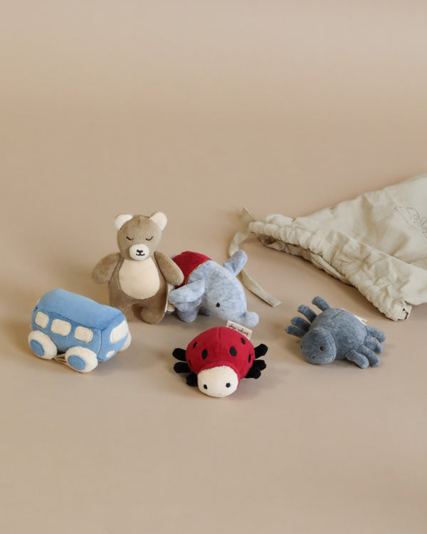 A group of velour toys is displayed against a beige background, including a teddy bear, a light blue bus, a red and black ladybug, a blue spider, and a blue dragon. A gray cotton bag is partially visible on the right side. This is the Konges Slojd | Five Piece Rattle Activity Set.