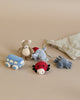 A group of velour toys is displayed against a beige background, including a teddy bear, a light blue bus, a red and black ladybug, a blue spider, and a blue dragon. A gray cotton bag is partially visible on the right side. This is the Konges Slojd | Five Piece Rattle Activity Set.