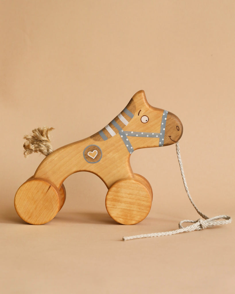 A Handmade Wooden Horse Pull Toy | Blue with painted details, a blue dotted harness, and a heart symbol on its side. This charming kids' toy with wheels features four circular wheels and a string attached to the front for pulling. The neutral beige background enhances its classic appeal.