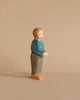 A handcrafted wooden figurine of a person stands against a beige background. Made by Ostheimer in Germany, the Ostheimer Father, Blue Sweater is painted with a blue top, gray pants, and beige shoes. Its plain, simple design conveys a minimalist aesthetic. The figurine has a neutral expression and is looking slightly forward.