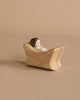 A small, minimalist wooden figurine of a person with dark hair is lying in a curved, boat-like wooden structure, set against a plain beige background. The handcrafted wooden toy's simple design and the natural wood tones create a serene, peaceful scene, perfect for imaginative play reminiscent of Ostheimer Child In Cradle.